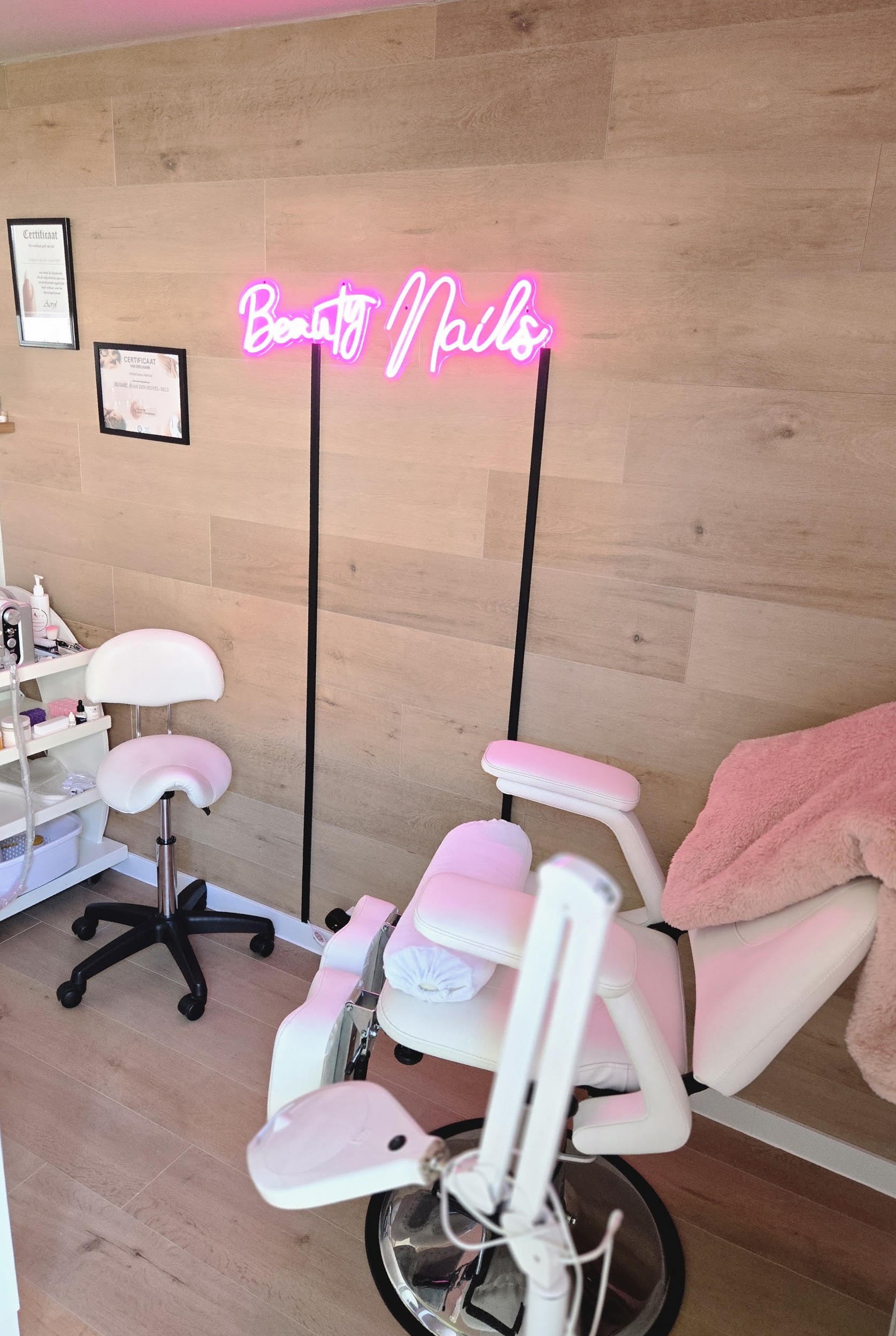 De salon: lichte houten wand, roze neonbord Beauty Nails en een witte pedicurestoel